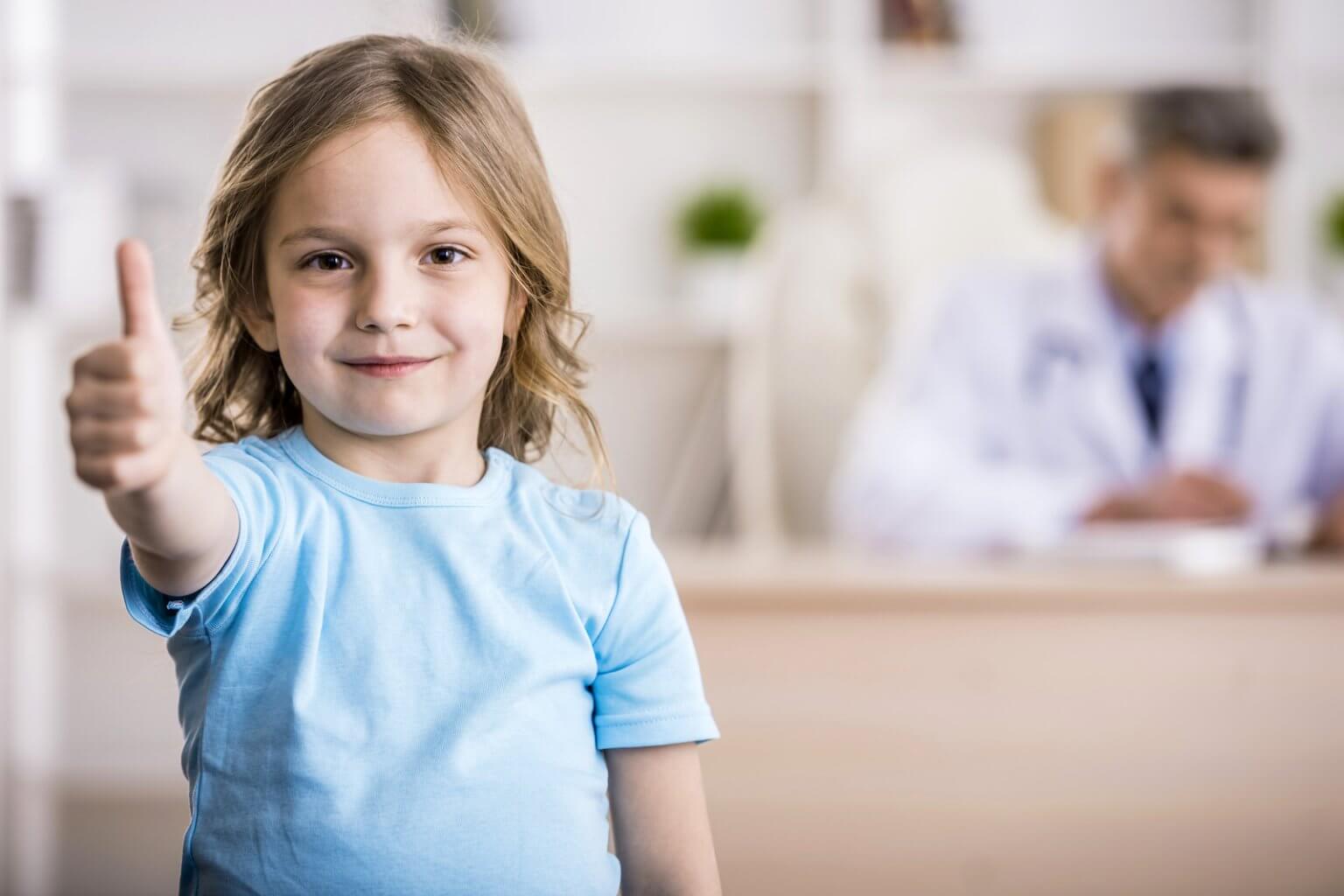 Cute little girl is showing thumb up. Doctor is sitting on background.